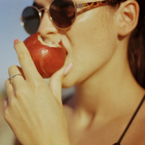 A woman with pink nails and a ring, wearing sunglasses, is about to bite into a red apple outdoors. Promote healthy habits with Eat more plants! (1-on-1 Health Coaching).
